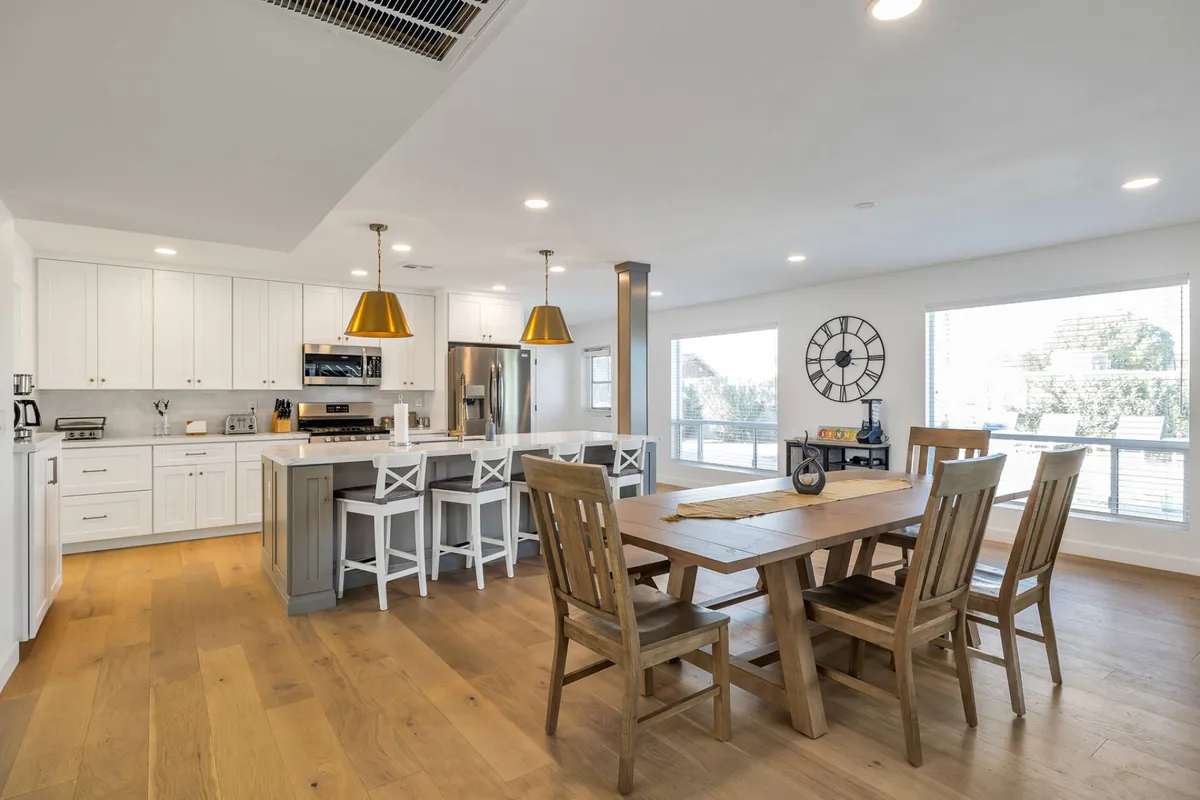 A bright and airy dining area is featured in this image, with a spacious table set for six and an adjoining kitchen island with barstools. Pendant lighting and large windows create a warm, welcoming mood, while the kitchen is fully visible, showcasing modern appliances and an inviting island perfect for gathering.