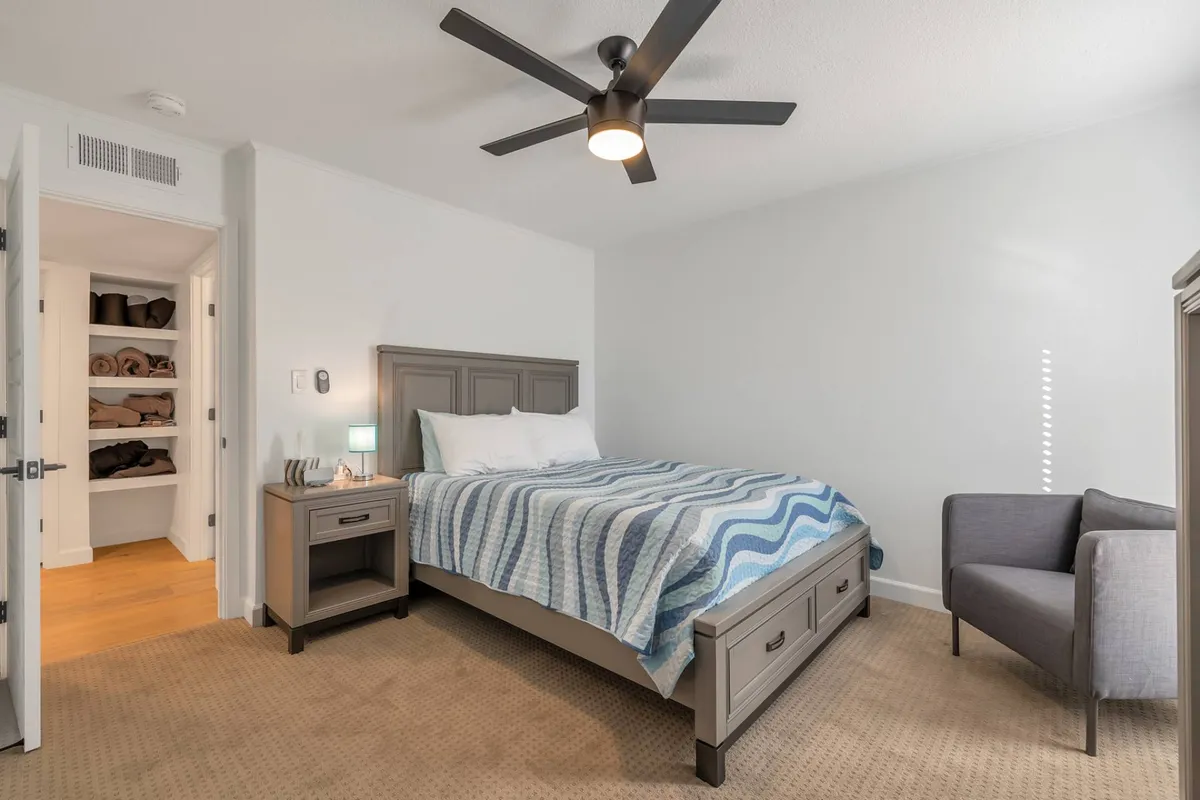 A calming guest bedroom featuring a bed with striped bedding, a cozy nightstand lamp, a seating area with a chair, and a closet neatly stocked with towels. The design is orderly and serene, promoting relaxation and comfort.