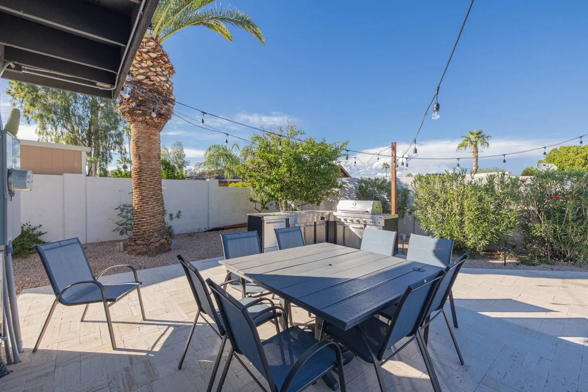 A vibrant outdoor dining setup is pictured, with a large dining table, a BBQ grill nearby, palm trees, and string lights overhead. The atmosphere is casual and festive, highlighting the alfresco dining and barbecue features.