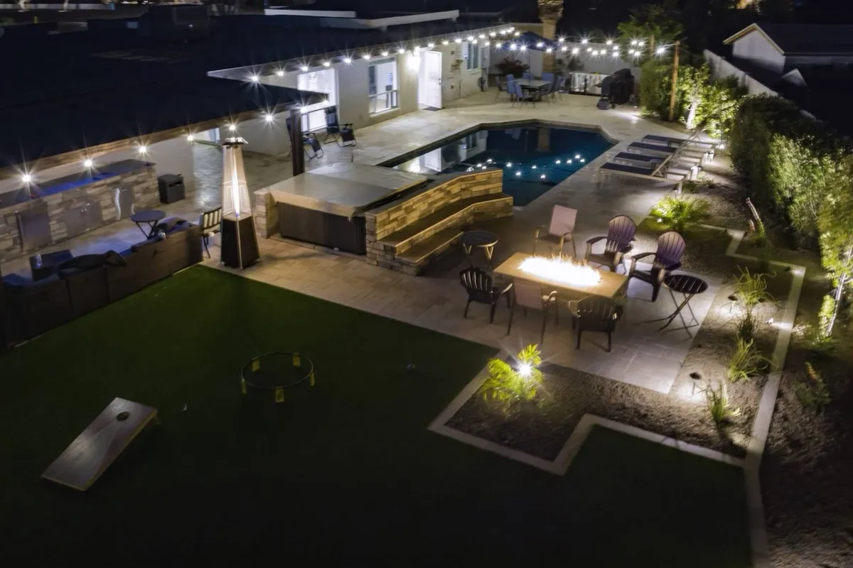 The image shows a stunning backyard oasis featuring a sparkling swimming pool, a cozy fire pit surrounded by comfortable outdoor seating, all beautifully illuminated by string lights. The space feels inviting and is perfect for evening gatherings, relaxation, and enjoying Phoenix's warm nights.