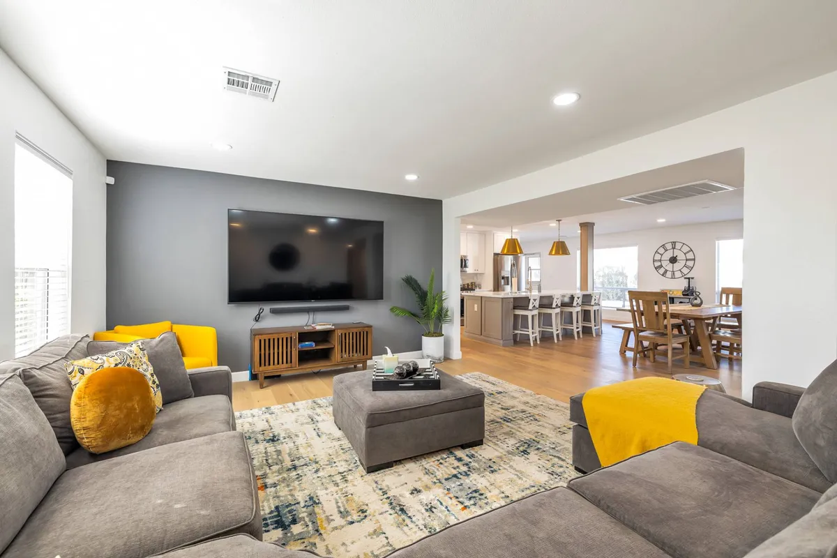 This image reveals a spacious open-plan living area where a plush sectional sofa faces a large television. The living zone flows seamlessly into a visible dining area, with ample natural light flooding the space. The coffee table and tasteful decor create a cozy and inviting environment that encourages relaxation and socializing.