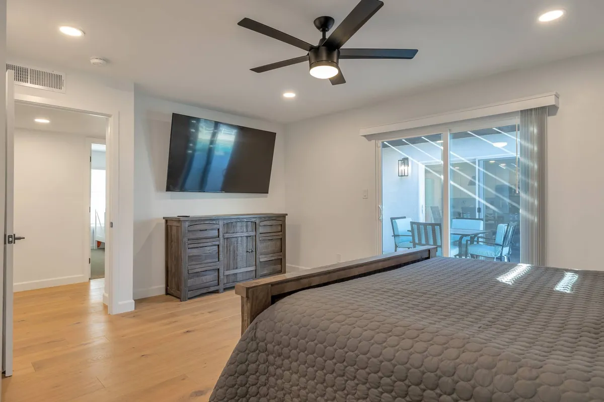 This image showcases a serene bedroom with a king bed, textured bedding, a large wall-mounted television, and a sliding glass door that opens directly to an outdoor space. A wooden dresser adds warmth while sunlight streams through the door, highlighting the bedroom's modern, restful style.
