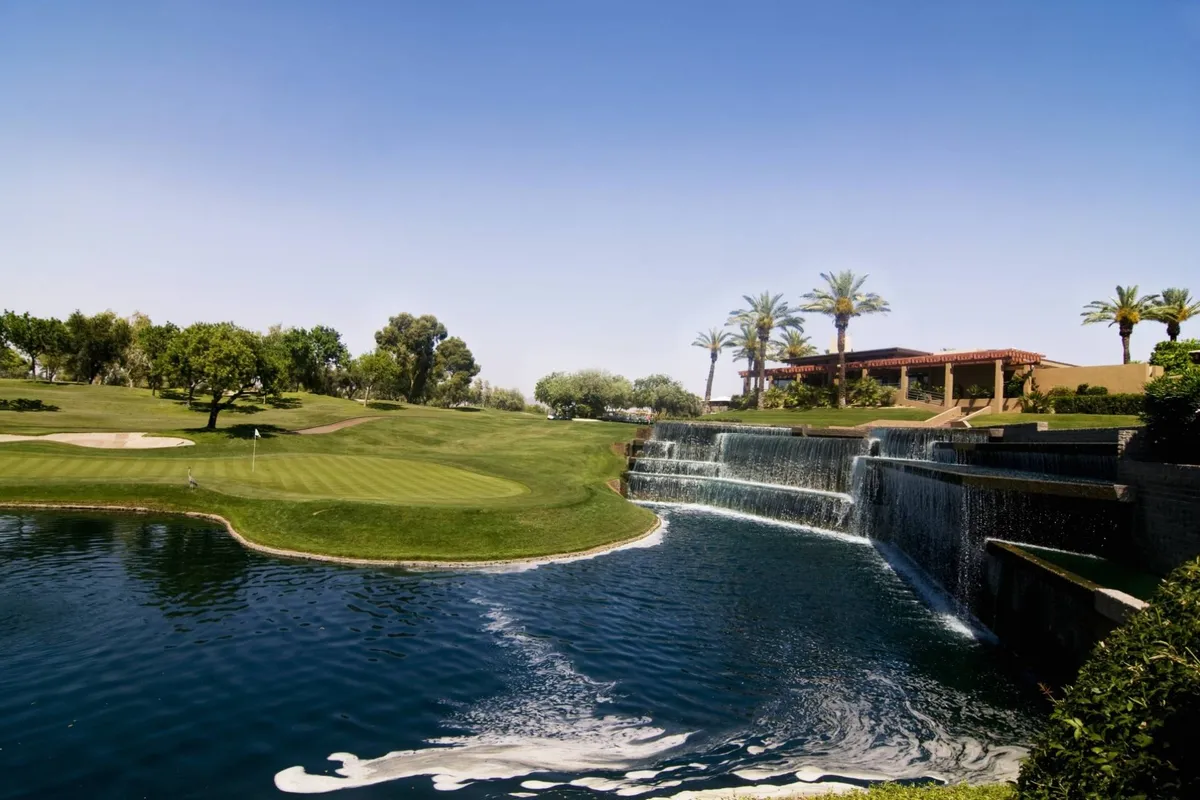 A lush, well-maintained golf course featuring vibrant green fairways, a picturesque waterfall surrounded by palm trees, and an inviting clubhouse in the distance. Golfers are visible on the course, enjoying a serene round, while others relax near the water or socialize around the clubhouse.